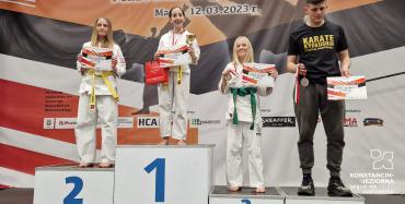 Trzy dziewczynki w białtcy strojach zawodniczek karate i jeden mężczyzna w czarnych dresach stoja na podiach. Na szyi maja medale, a w dłoniach dyplomy.