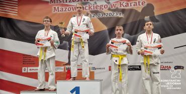 Czterech chłpców w białych strojach zawodników karate stoi na podium z dyplomami w dłoniach i pucharem. 