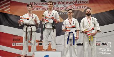 Czterech młodych mężczyzn sttoi na podium. Są ubrani w białe stroje sportow dla zawodników karate, na szyi maja medale, a w dłonich dyplomy.