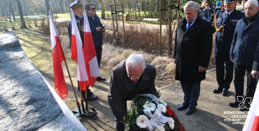 Starszy mężczyzna składa wieniec pod kamieniem. Obok niego są umieszczone polskie flagi.