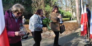 Trzy starsze kobiety idą alejką parkową. Trzymaja w dłoniach kwiaty i znicze. 