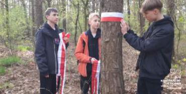 Chłopiec przywiązuje do pnia drzewa w lesie biało-czerwona tasiemkę. Dwóch pozostałych z tasiemkami w dłoniach stoi obok.