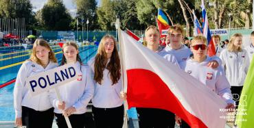 Trzy dziewczyny trzymają tabliczkę z napisem „Poland”. Obok stoi trzech chłopców i trzymają biało-czerwona flagę. Za nimi basen i drzewa