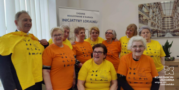 Dziewieć starszych kobiet i jeden starszy mężczyzna w żółtych i pomarańczowych koszulkach. Za nimi baner z napisem: zadanie realizowane w ramach inicjatywy lokalnej.