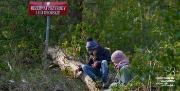 Dwójka dzieci siedzi na pniu, wokół nich zielone drzewa i krzaki. W drugim planie czerwona tabliczka z białym napiem: Rezerwat Przyrody Łegi Oborskie.