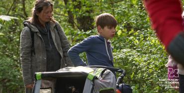 Kobieta i chłopiec z wózkiem spacerowym dla małego dziecka. Za nimi drzewa z zielonymi listkami.
