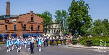 Rondo po którym idzie w trzech szeregach orkiestra strażacka oraz dziewczynki w biało-błękitnych sukienkach. Za nimi budynek z cegły i z wyokim kominem.