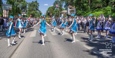 Dziewczyny w błękitno-białych sukienkach, z błękitnymi czapeczkami na głowach sa w kręgu na ulicy; maja uniesiona prawą nogęoraz lekko rozłozone na boki ręce. Za nimi stoi tłum ludzi.