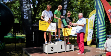 Trzej młodzi męzczyźni stoja na podium. Czwarty, starszy mężczyzna stoi przed nimi.