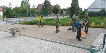 Grupa młodych ludzi w mundurach harcerskich. Stoją na chodniku, są pochyleni, a w dłoniach mają miotły.