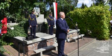 Starszy mężczyzna w garniturze stoi obok nagrobka, przed nim jest statyw z mikrofonem.