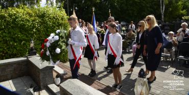 Diw dziewczynki i jedench chłopiec w białych bluzkach. Chłopak trzyma w ręce wieniec z biało-czerwonych kwiatów.