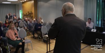 Sala konferencyjna. Osoby siedzą na krzesłach i patrzą na mężczyznę z mikrofonem w ręce.