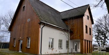 Bydynek z dachem spadzistym widoczny od narożnika, jedna ściana pokryta drewniem, na ścianie po prawej stronie widoczna nadbudówka  pokryta drewnem i podparta kolumnami