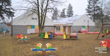 Na pierwszym planie kolorowe urządzenia placu zabaw - huśtawki, bujaczki i inne, z nimi widoczny obiekt tworzony przez dwa identyczne dudynki ze spadzistymi dachami połączonymi łącznikiem, na ścianach panele kolorowe