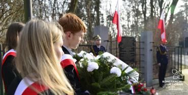 Młodzi ludzie z kwiatami. W tle tablica zawieszona na ogrodzeniu, a obok niej zawieszone biało-czerwone flagi.