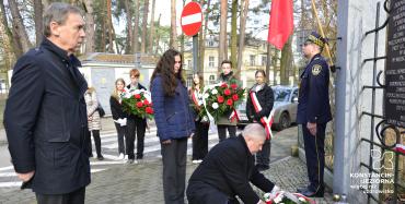 Starszy mężczyzna kuca przy ogrodzeniu i poprawia biało-czerwone kwiaty.