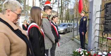 Na wprost tablicy zawieszonej na ogrodzeniu stoi z pochyloną głową kobieta oraz dwie dziewczyny i chłopiec.