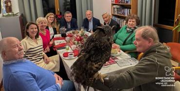 Uczestnicy świątecznego spotkania stoją wokół stołu w sali urzędu, słuchając opowieści o sowach, które znajdują się na środku pomieszczenia.