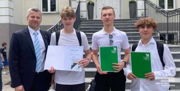 Zastępca burmistrza gminy oraz trzej członkowie zwycięskiej drużyny pozują do pamiątkowego zdjęcia przed budynkiem.
