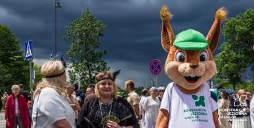 Uczestniczki Pikniku Retro rozmawiają z miejską maskotką wiewiórki na tle ciemnych chmur i zgromadzonych mieszkańców.