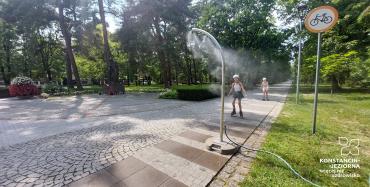 Dzieci na rolkach przejeżdżają przez mgiełkę wodną rozpylaną przez słupek zraszający.