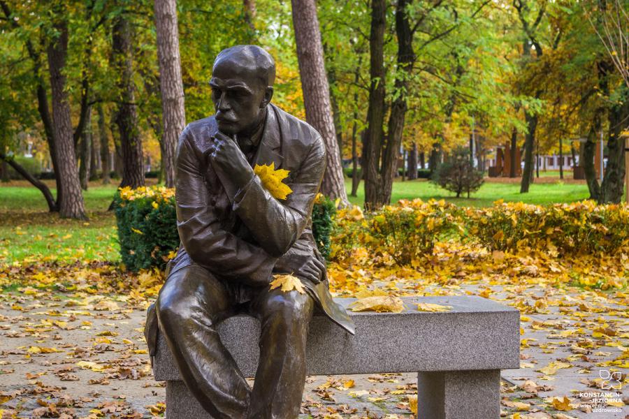 A cast metal monument depicting a middle-aged man sitting on a bench, with autumn trees around it 