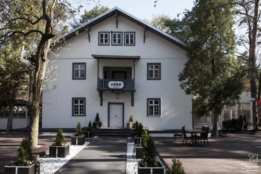 The front of the two-story building with a sloping roof, above the entrance, with the date 1899, around a tree. 