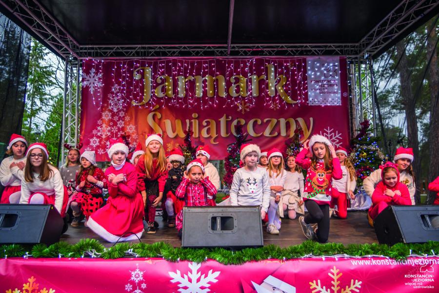 An open-air scene with a roof, with a group of children in Santa's Christmas costumes. with them a visible banner with the name of the Christmas Fair
