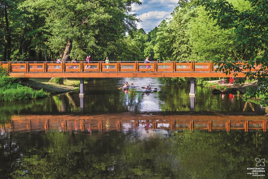 A wooden bridge stretched in the park by the river on which people in canoes are swimming, people walk and cycle on the bridge 