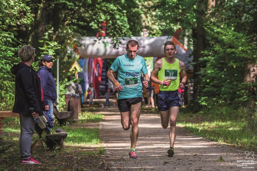 Two running men in sports clothes on a forest path with viewers standing to the side 