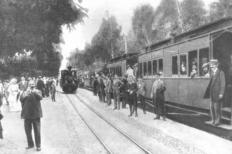 Old black-white photograph showing a railway station with a train and people getting on