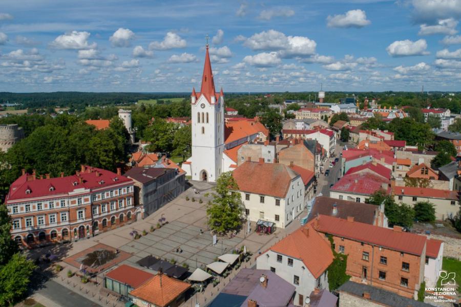 A view from the top of the market square old town, centrally visible church with a high tower, buildings around old houses with red tiles, in the background a lot of greenery outside the city 