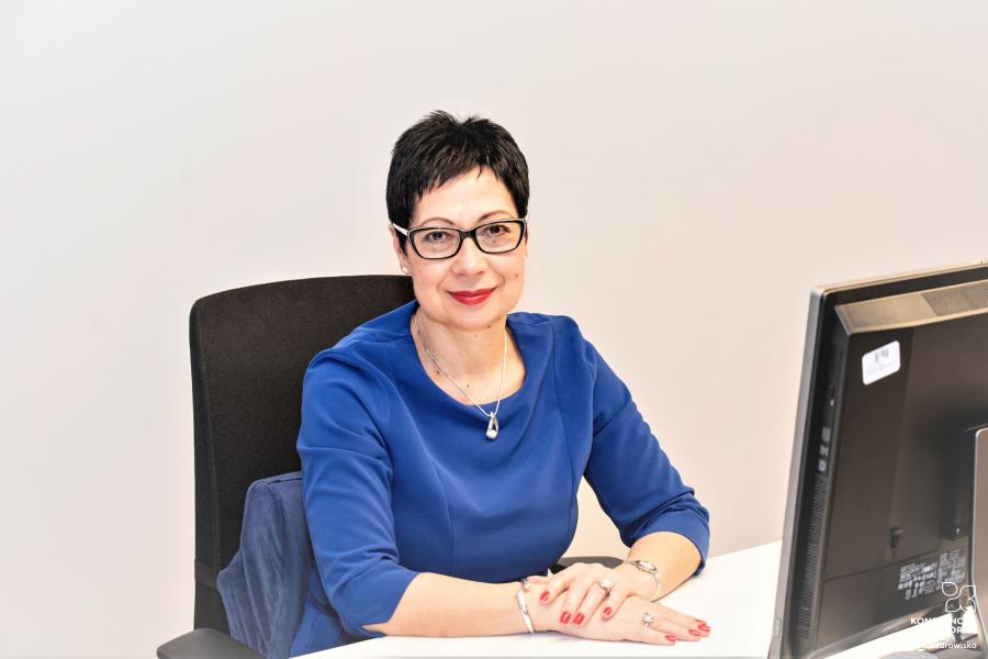 Uśmiechnięta sekretarz gminy Bożena Pindelska siedzi przy biurku, przed nią stoi monitor od komputera.