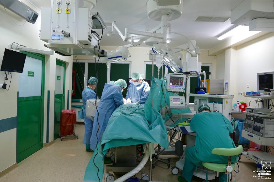 The operating room, several people in surgical attire are standing around the table, lamps hang above them 