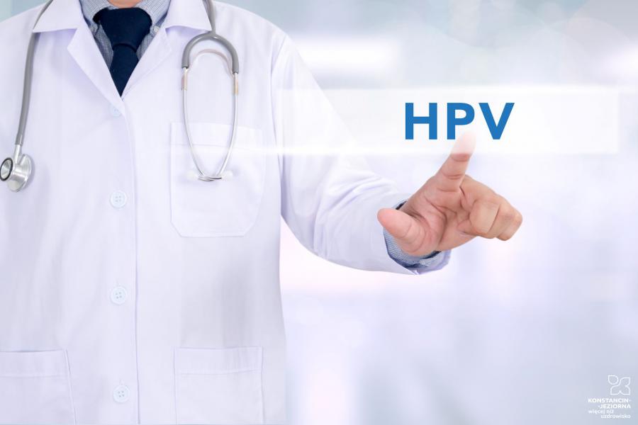 Lekarz ubrana w biały fartuch, na szyi ma słuchawki medyczne, lewą ręką wskazuje na tabliczkę z napisem HPV. 