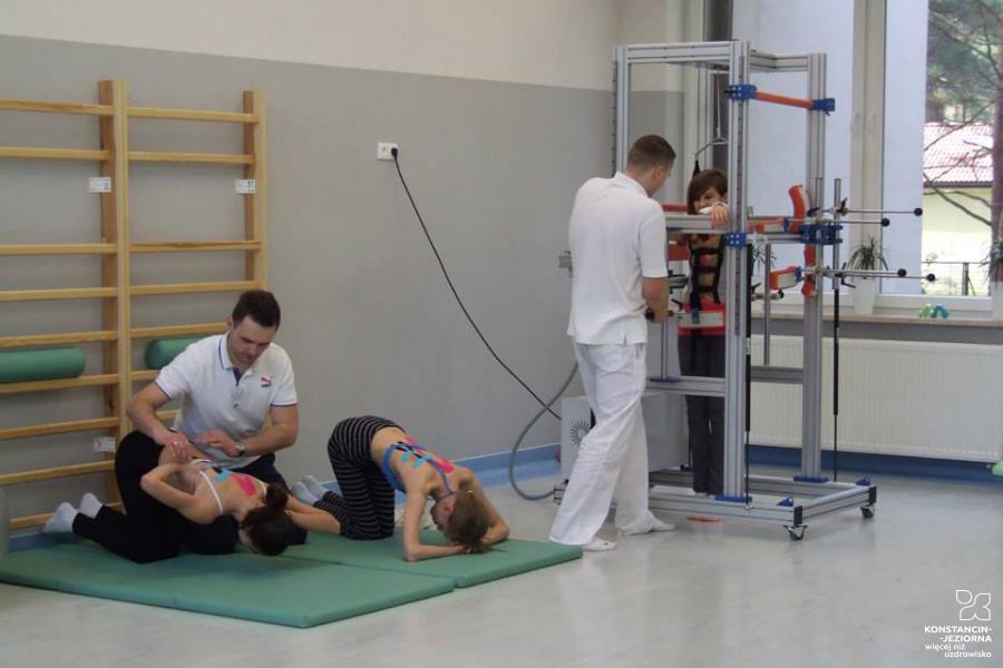Rehabilitation room, a ladder on the wall on the left, a patient is lying on the mattress underneath it and a physiotherapist is kneeling next to him, another patient is exercising in the device on the right 