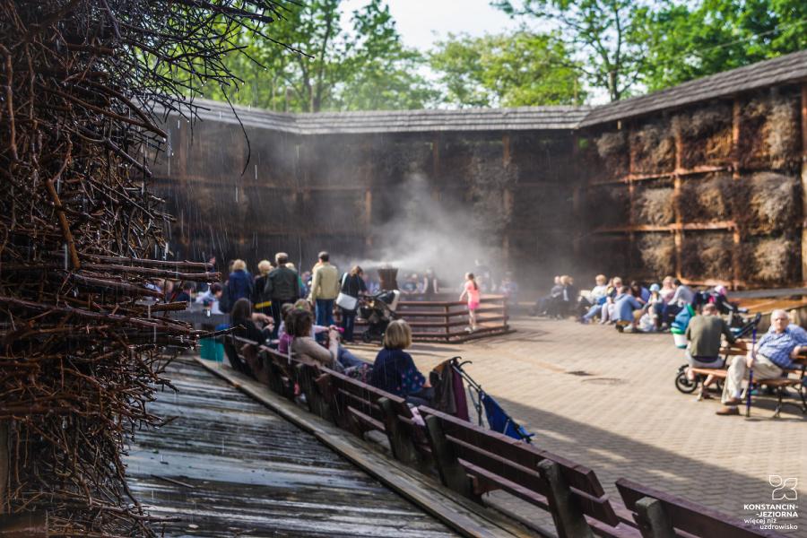 The interior of the open building is surrounded by walls filled with branches, people sit around on benches, water vapor rises 