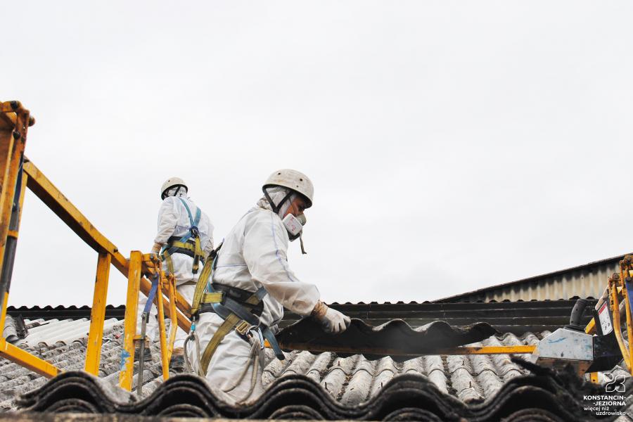 Na zdjęciu dwóch pracowników specjalistycznej firmy do usuwania azbestu z dachów. Są ubrani w białe kombinezony, maski i kaski. Demontują płyty azbestowe z dachu budynku.