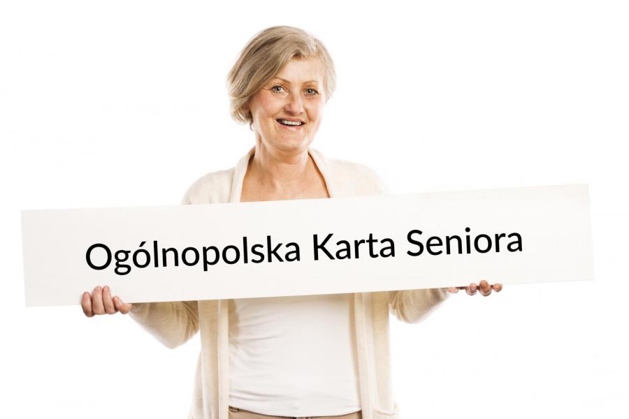 Na zdjęciu uśmiechnięta seniorka, która w rękach trzyma dużą kartę papieru, na której znajduje się napis - Ogólnopolska Karta Seniora. 