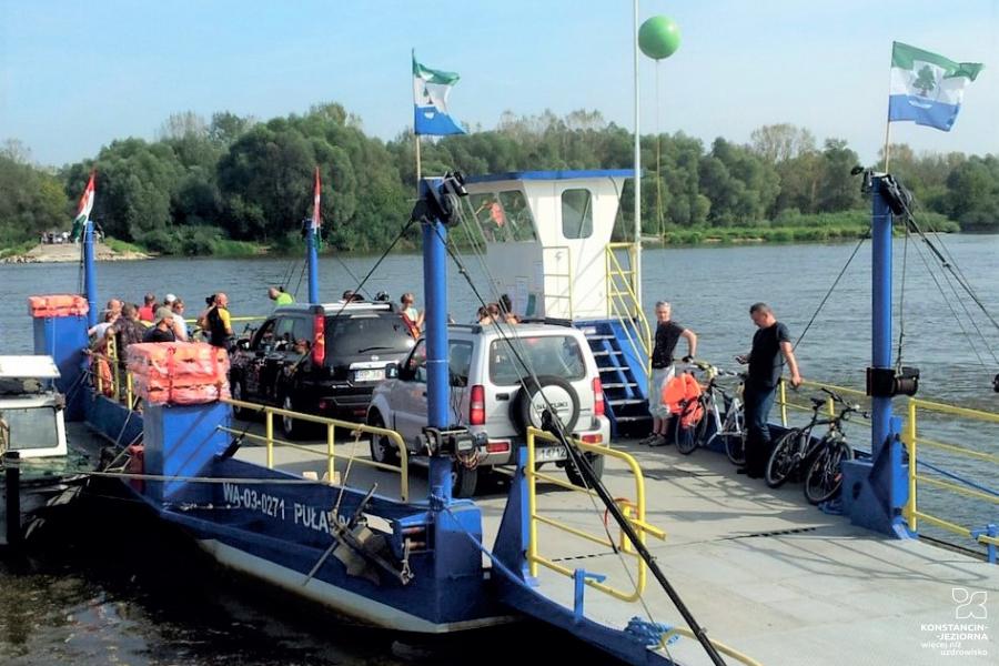 Fragment rzeki Wisły z promem, na którym stoją dwa samochody oraz kilka osób. W powietrzu  powiewają flagi gminy Konstancin-Jeziorna