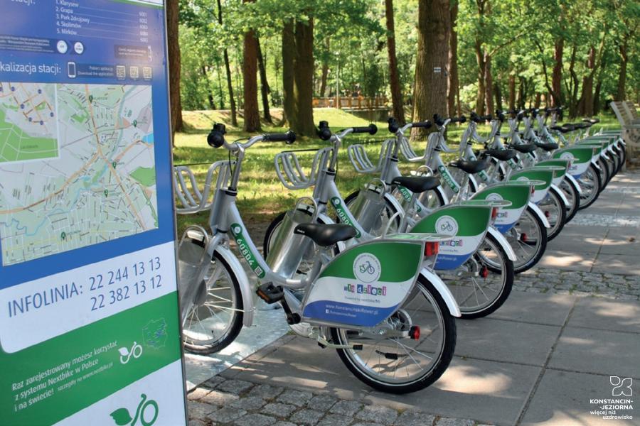 Duży stojak na rowery, na którym obok siebie stoi około 20 rowerów do wypożyczenia. Stojak stoi przy ścieżce spacerowej w Parku Zdrojowym. 