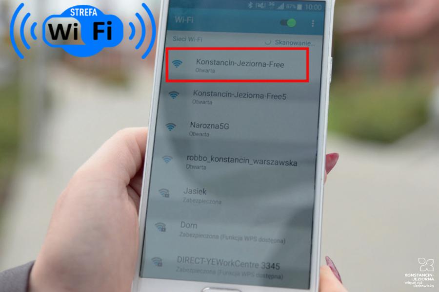 Ręka trzymająca smartfona, na ekranie lista dostępnych sieci wifi, czerwoną ramką zaznaczona sieć o nazwie Konstancin-Jeziorna Free, w górnym lewym roku napis strefa wifi.