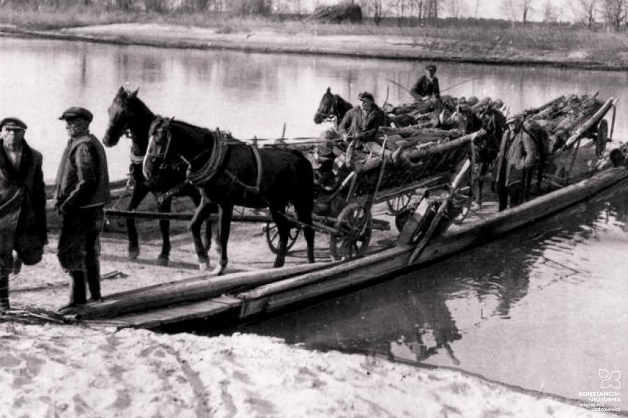 Czarno-biała fotografia. W głównej części duża drewniana tratwa przycumowana do brzegu Wisły. Na tratwie znajdują się 3 konie, wozy drewniane i 5 pasażerów. Wozy załadowane są drewnem.