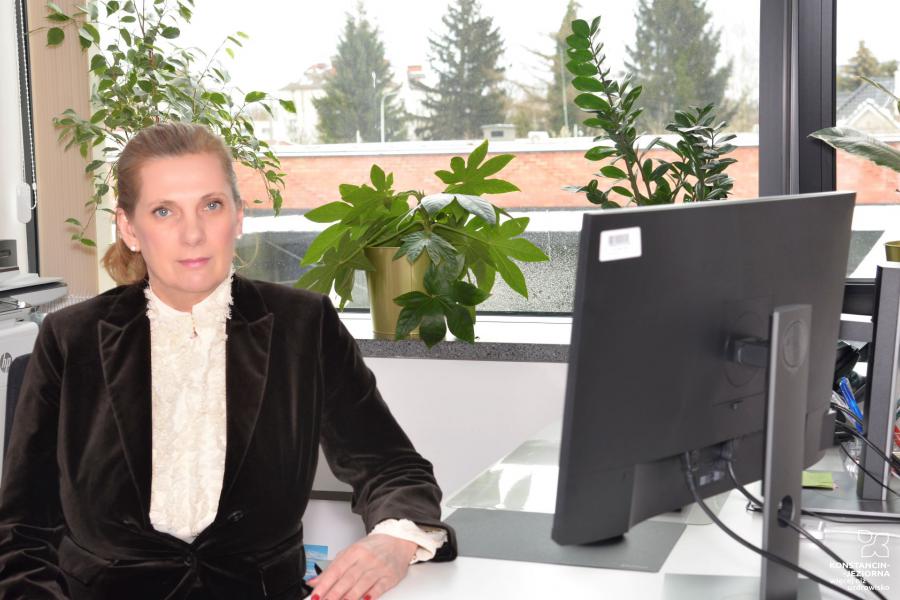 Uśmiechnięta kobieta siedzi przy biurku, przed nią stoi monitor od komputera. Ma długie blond włosy. Ubrana jest w kolorową sukienkę. 