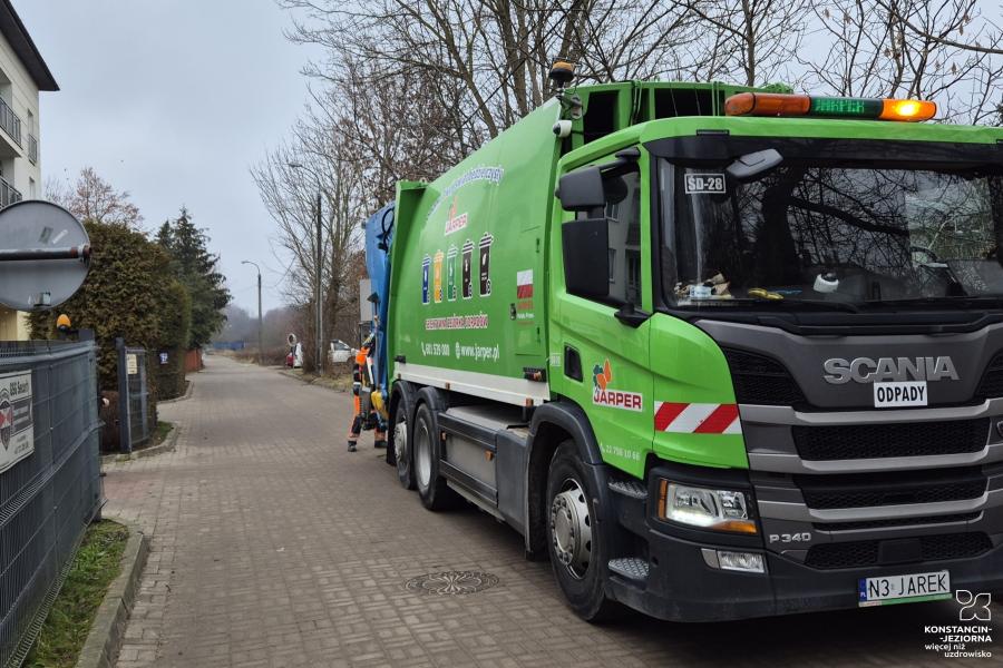 zielona śmieciarka marki Scania należąca do firmy Jarper, zaparkowana na osiedlowej ulicy, podczas gdy pracownik w pomarańczowym uniformie obsługuje kontenery na odpady.