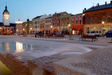 Widok na rynek miejski, z zabytkową zabudową, po prawej stronie szereg kamieniczek, na lewo fragment budowli sakralnej z wysoką wieżą. W centralnej części znajduje się duża fontanna.