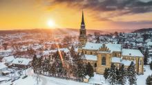 Widok z góry na pokryte śniegiem miasto. W centralnej części, pośród drzew iglastych znajduje się kościół z wysoką wieżą. W tle panorama gór oraz wielobarwne niebo  z zachodzącym słońcem.