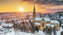 Widok z góry na pokryte śniegiem miasto. W centralnej części, pośród drzew iglastych znajduje się kościół z wysoką wieżą. W tle panorama gór oraz wielobarwne niebo  z zachodzącym słońcem.