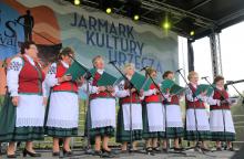 Zdjęcie przedstawia zespół Łurzycanki występujący na scenie podczas Jarmarku Kultury Urzecza. Osiem pań, ubranych jest w tradycyjne stroje z regionu. W rękach trzymają śpiewniki. 